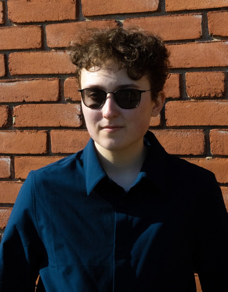 A headshot of a man with short, curly hair in a dark blue shirt and sunglasses.