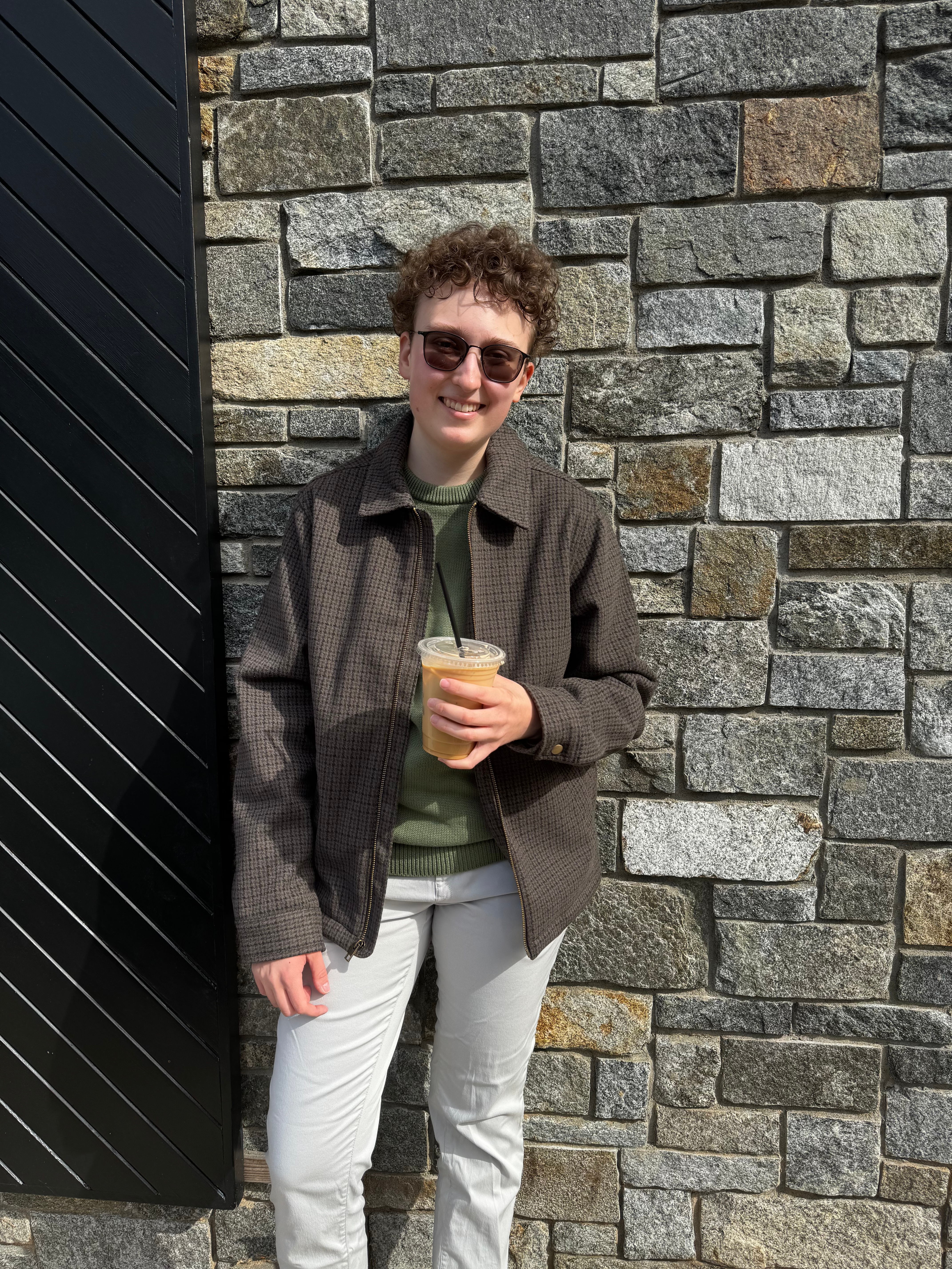 Taylor leans up against a brick wall and poses with an iced coffee.